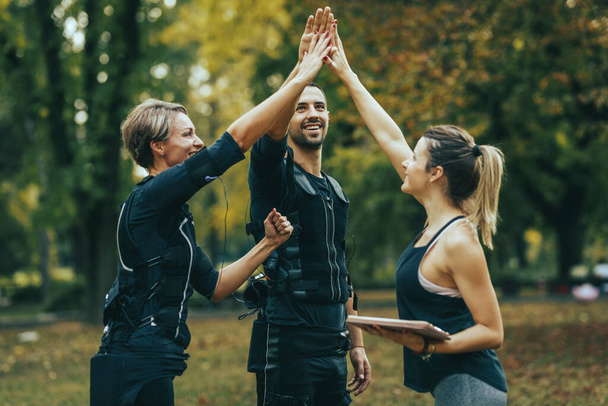 Ένα ταιριαστό ζευγάρι που γιορτάζει με γυναίκα προσωπικό γυμναστή μετά από επιτυχή προπόνηση στο πάρκο, ντυμένο με μαύρη στολή με ηλεκτρονικό προσομοιωτή EMS για την τόνωση των μυών τους. - Φωτογραφία, εικόνα