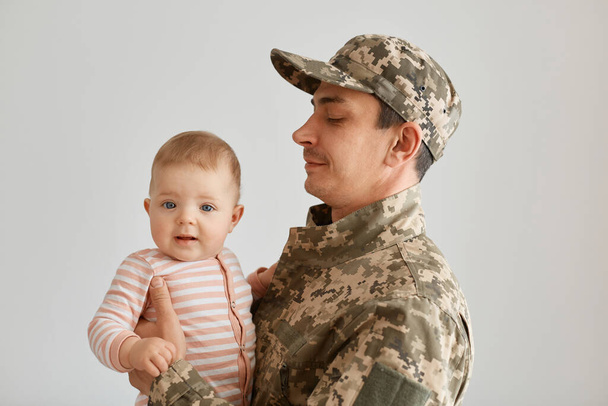 Side view of young adult military father came from the war or the exercises and holding hid cute baby girl wearing striped sleeper, armyman wearing camouflage uniform and cap. - 写真・画像