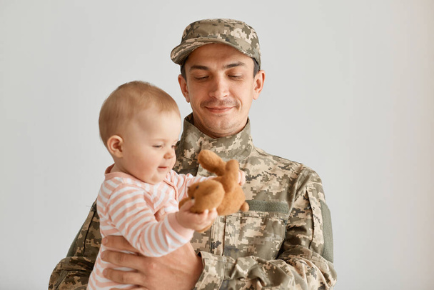 Positive young adult military man wearing camouflage uniform and cap, standing with his little infant daughter in hands and looking kid plaing with soft toy. - Fotoğraf, Görsel