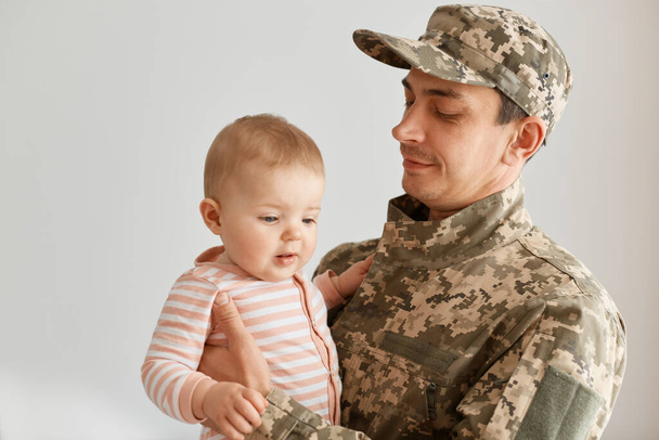 Indoor shot of military father came from the war or the exercises and posing with his little toddler daughtr in striped sleeper, posing at home after returning home. - Foto, immagini