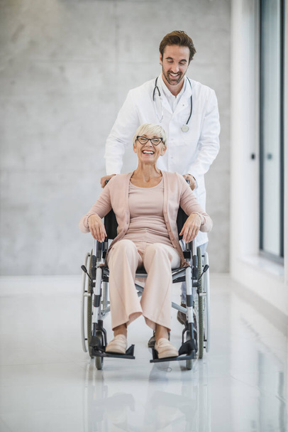 Πυροβολισμός ενός νεαρού γιατρού που φροντίζει μια ηλικιωμένη γυναίκα ασθενή σε αναπηρική καρέκλα. - Φωτογραφία, εικόνα