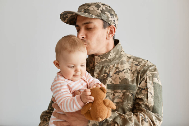 Indoor shot military man wearing camouflage uniform and cap, standing with his little infant daughter in hands, father missing his child and kissing baby, expressing love. - Фото, изображение