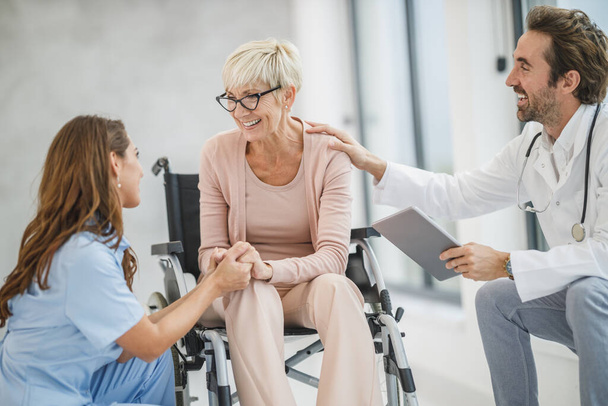 Medico di successo e giovane infermiera che parlano con la loro paziente anziana sulla sedia a rotelle. - Foto, immagini