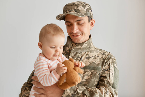 Indoor shot of military father came from the war or the exercises and cheerfully holding baby with soft toy, being happy to return home and see hid kid. - Fotó, kép