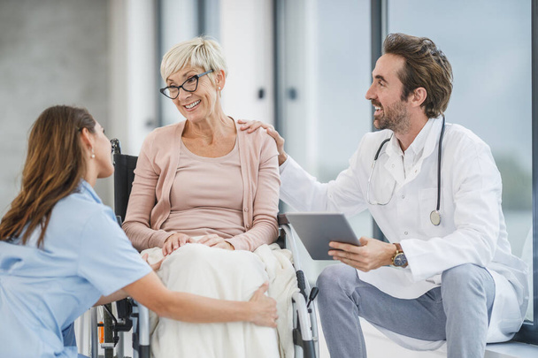 Успешный врач и молодая медсестра разговаривают со своей старшей пациенткой в инвалидной коляске. - Фото, изображение