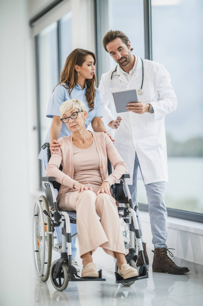 Επιτυχημένος γιατρός και νεαρή νοσοκόμα συνομιλούν με την ηλικιωμένη γυναίκα ασθενή με αναπηρική καρέκλα. - Φωτογραφία, εικόνα