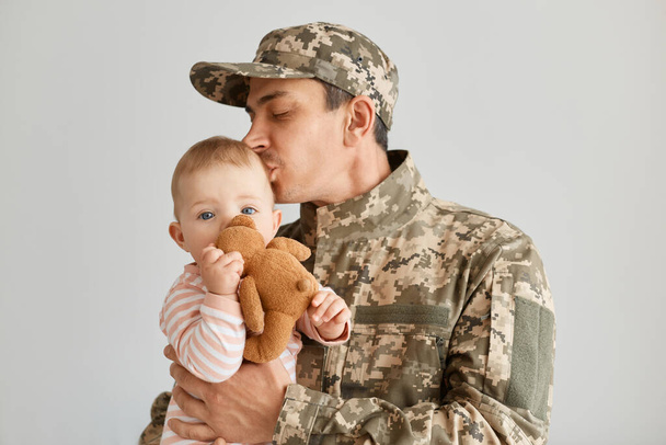 Portrait of young adult military father came from the war or the exercises and kissing his toddler daughter, baby holding soft toy, male wearing uniform and cap, - Φωτογραφία, εικόνα