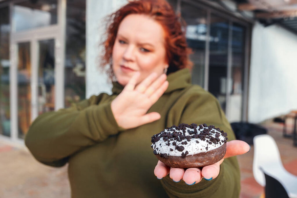 A blurry portrait of a well-fed woman holding a chocolate donut in the palm of her hand. The concept of diet and weight loss. - Photo, Image