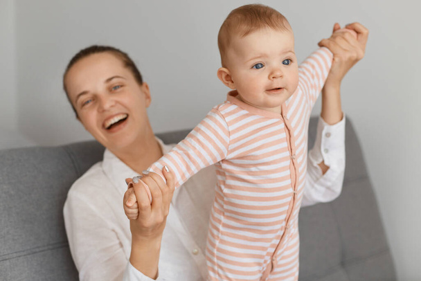 Portrait of Caucasian mother playing with her infant baby, family spending time together, woman with hair bun laughing happily while sitting with her toddler daughter on sofa. - Foto, imagen