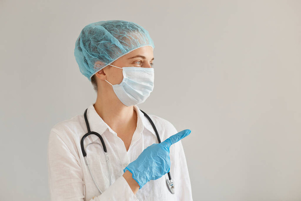 Horizontal shot of woman doctor wearing medical cap, gloves, surgical mask and gown, showing mockup for promotional text or your advertisement with fore finger. - Fotó, kép