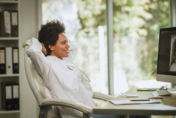 Aufnahme einer afrikanischen Ärztin, die allein am Schreibtisch sitzt und sich entspannt, nachdem sie während der COVID-19-Pandemie Röntgenaufnahmen am Computer in ihrem Sprechzimmer gemacht hat. - Foto, Bild