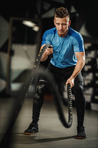 Aufnahme eines muskulösen Kerls in Sportbekleidung, der im Fitnessstudio Übungen mit Seilen macht. - Foto, Bild