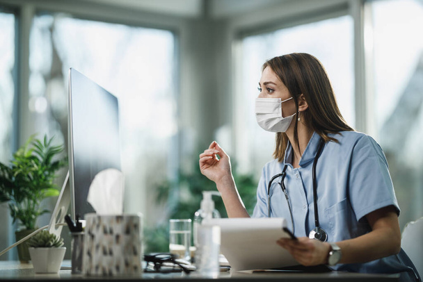 Atrakcyjna młoda pielęgniarka z maską ochronną przy użyciu komputera podczas pracy w szpitalu. - Zdjęcie, obraz