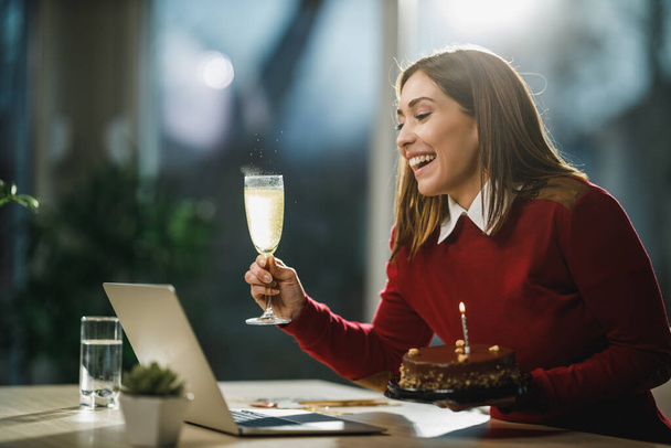 Lachende jonge vrouw met een verjaardagstaart en een glas champagne en hebben online verjaardagsfeest met vrienden tijdens coronavirus pandemie. - Foto, afbeelding
