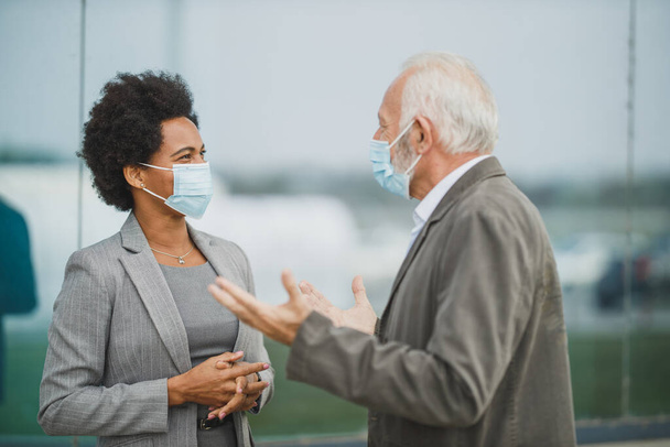 Strzał odnoszącej sukcesy czarnej bizneswoman i jej starszego kolegi płci męskiej za pomocą maski ochronnej podczas dyskusji przed budynkiem biurowym podczas pandemii COVID-19. - Zdjęcie, obraz