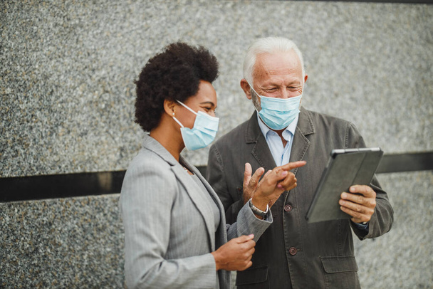 Aufnahme eines erfolgreichen Geschäftsmannes und seiner schwarzen Kollegin mit Schutzmaske, die ein digitales Tablet benutzt und eine Diskussion führt, während sie während der COVID-19-Pandemie vor einer Wand eines Firmengebäudes steht. - Foto, Bild