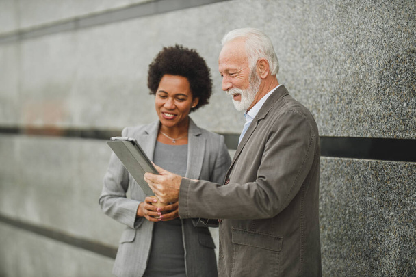 Aufnahme zweier erfolgreicher multiethnischer Geschäftsleute, die ein digitales Tablet benutzen und eine Diskussion führen, während sie vor einer Wand eines Firmengebäudes stehen. - Foto, Bild