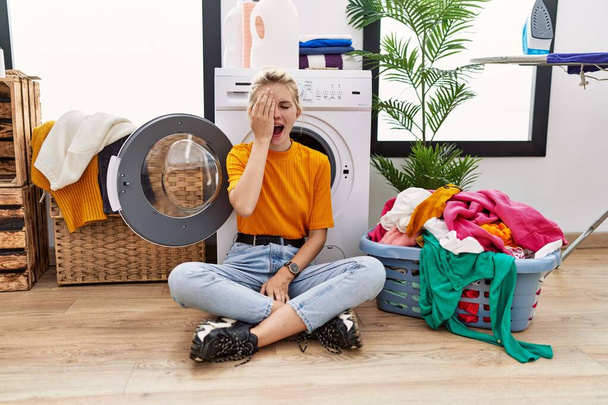 Young blonde woman doing laundry sitting by washing machine yawning tired covering half face, eye and mouth with hand. face hurts in pain.  - Foto, Bild