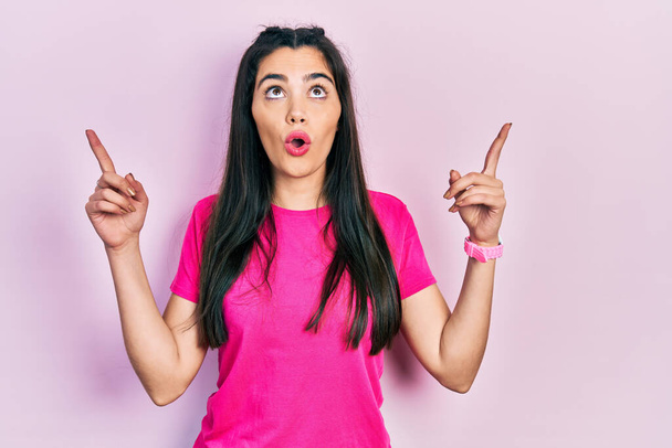 Молода іспаномовна дівчина в повсякденній рожевій сорочці здивована і здивована, дивлячись вгору і вказуючи пальцями і підняті руки
.  - Фото, зображення