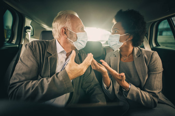 Shot of two worried multi-ethnic business people with protective mask having a discussion about work while sitting in the backseat of a car on their morning commute during COVID-19 pandemic. - Photo, Image