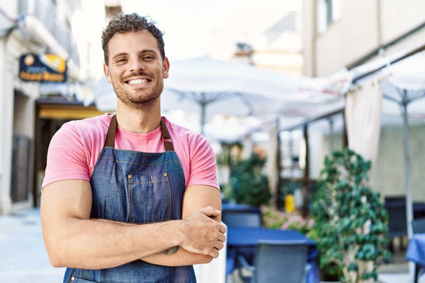 Jonge Spaanse ober draagt schort met gekruiste armen in de koffieshop. - Foto, afbeelding