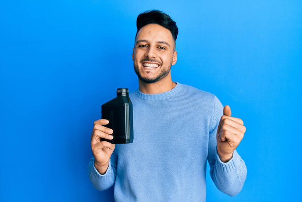Young arab man holding motor oil bottle screaming proud, celebrating victory and success very excited with raised arm  - Foto, Imagen