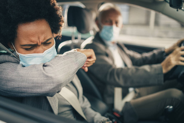 COVID-19パンデミック時に車で移動中に肘で保護マスク咳を持つ成功した黒人ビジネス女性. - 写真・画像