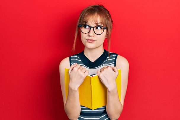 Κοκκινομάλλα νεαρή γυναίκα διαβάζει ένα βιβλίο φορώντας γυαλιά χαμογελώντας κοιτώντας στο πλάι και κοιτάζοντας αλλού σκεπτόμενη.  - Φωτογραφία, εικόνα