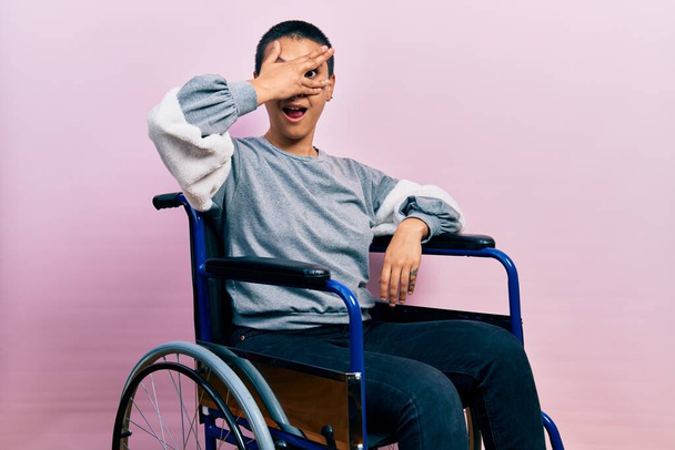 Beautiful hispanic woman with short hair sitting on wheelchair peeking in shock covering face and eyes with hand, looking through fingers with embarrassed expression.  - Fotoğraf, Görsel
