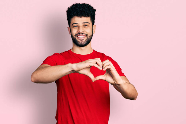Young arab man with beard wearing casual red t shirt smiling in love doing heart symbol shape with hands. romantic concept.  - Foto, imagen