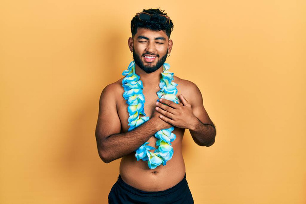 Arabische man met baard in badpak en Hawaïaanse lei lachend met handen op de borst met gesloten ogen en dankbaar gebaar op gezicht. Gezondheidsconcept.  - Foto, afbeelding