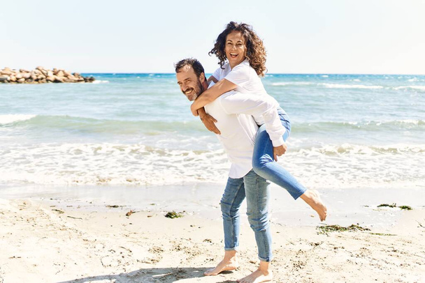 Latynos w średnim wieku uśmiechający się szczęśliwy trzymając kobietę na plecach na plaży. - Zdjęcie, obraz