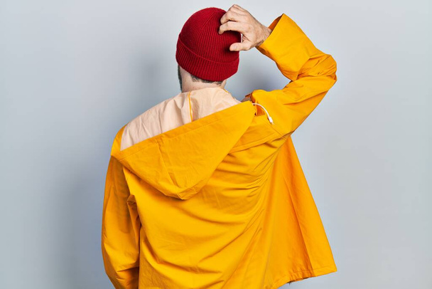 Homme caucasien avec barbe portant imperméable jaune vers l'arrière pensant au doute avec la main sur la tête  - Photo, image