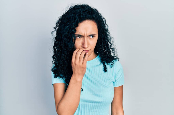Молода іспаномовна жінка з кучерявим волоссям в повсякденній синій сорочці виглядає напруженою і нервує руками на роті, кусаючи нігті. проблема тривоги
.  - Фото, зображення