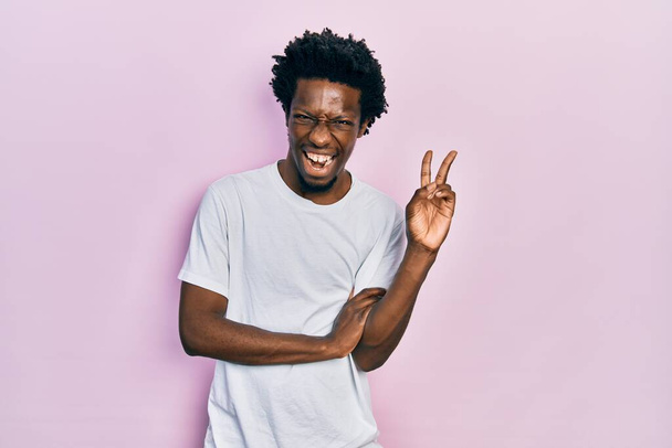 Ein junger afrikanisch-amerikanischer Mann in einem lässigen weißen T-Shirt lächelt mit glücklichem Gesicht und zwinkert in die Kamera, als er ein Siegeszeichen macht. Nummer zwei.  - Foto, Bild