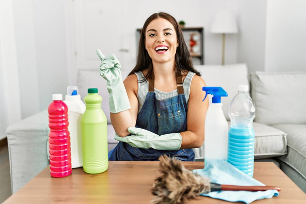 Young brunette woman wearing cleaner apron and gloves cleaning at home with a big smile on face, pointing with hand and finger to the side looking at the camera.  - Foto, imagen