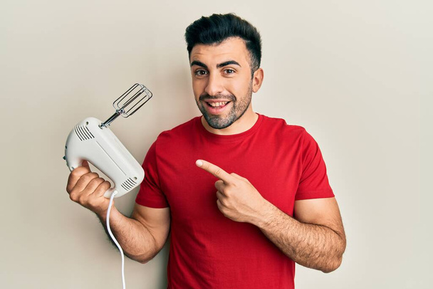 Young hispanic man holding food processor mixer machine smiling happy pointing with hand and finger  - Zdjęcie, obraz