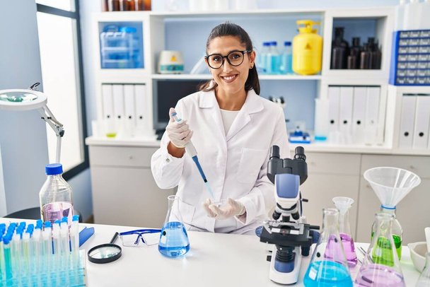 Junge hispanische Frau in Wissenschaftleruniform mit Pipette im Labor - Foto, Bild