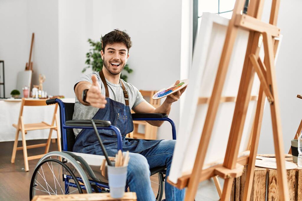 Молодий іспаномовний чоловік сидить на інвалідному візку в художній студії, посміхаючись доброзичливо пропонуючи рукостискання як привітання та привітання. успішний бізнес
.  - Фото, зображення