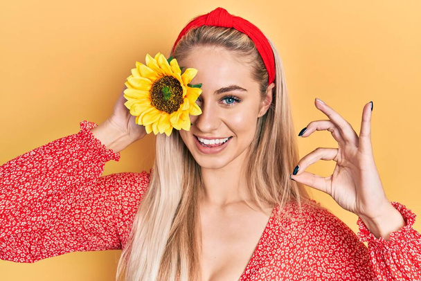 Mladá krásná běloška drží žluté slunečnice přes oko dělá ok znamení s prsty, usmívá přátelské gesto vynikající symbol  - Fotografie, Obrázek