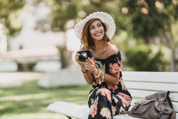 Μια ελκυστική νεαρή γυναίκα με ψηφιακή φωτογραφική μηχανή απολαμβάνοντας καλοκαιρινές διακοπές, ενώ κάθεται σε ένα παγκάκι στο πάρκο. - Φωτογραφία, εικόνα