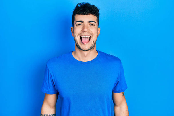Fiatal spanyol férfi alkalmi kék pólóban, kidugja a nyelvét vidáman vicces arckifejezéssel. érzelem koncepció.  - Fotó, kép