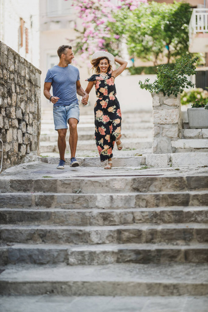 Χαρούμενο ζευγάρι ερωτευμένο να διασκεδάζει και να απολαμβάνει τις καλοκαιρινές διακοπές ενώ τρέχει κάτω από τις σκάλες στη μεσογειακή πόλη. - Φωτογραφία, εικόνα