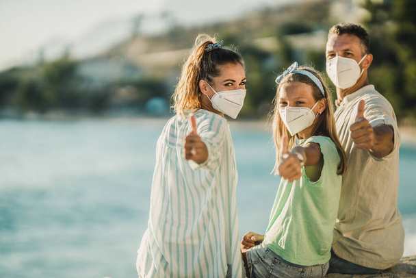 Ein lächelndes Teenager-Mädchen und ihre Eltern mit schützender N95-Maske, die Zeit am Meer verbringen und während der Coronapandemie Daumen nach oben zeigen.  - Foto, Bild