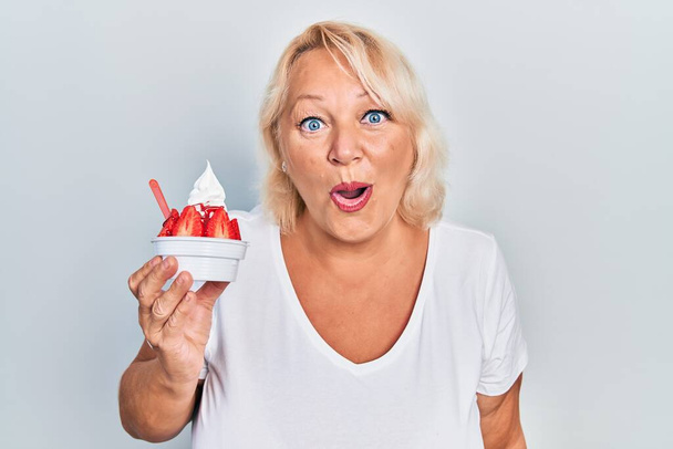 Středního věku blondýny žena jíst jahodovou zmrzlinu strach a ohromen s otevřenými ústy pro překvapení, nevěřící tvář  - Fotografie, Obrázek