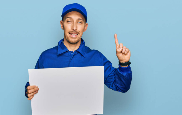 Φαλακρός άντρας με γενειάδα φορώντας στολή οικοδόμου κρατώντας άδειο πανό έκπληκτος με μια ιδέα ή ερώτηση δείχνοντας δάχτυλο με χαρούμενο πρόσωπο, νούμερο ένα  - Φωτογραφία, εικόνα