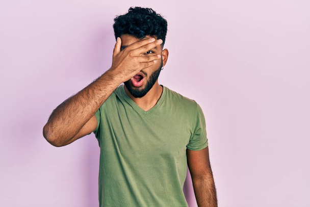 Арабский мужчина с бородой в непринужденной зеленой футболке подглядывает в шокирующее лицо и глаза рукой, испуганно глядя сквозь пальцы  - Фото, изображение