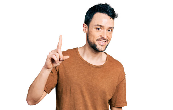 Homem hispânico com barba vestindo camiseta casual sorrindo com uma ideia ou pergunta apontando o dedo para cima com o rosto feliz, número um  - Foto, Imagem