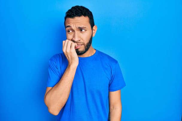 Sakallı, günlük mavi tişörtlü İspanyol bir adam gergin görünüyor ve elleri ağızdan tırnak yiyor. anksiyete.  - Fotoğraf, Görsel