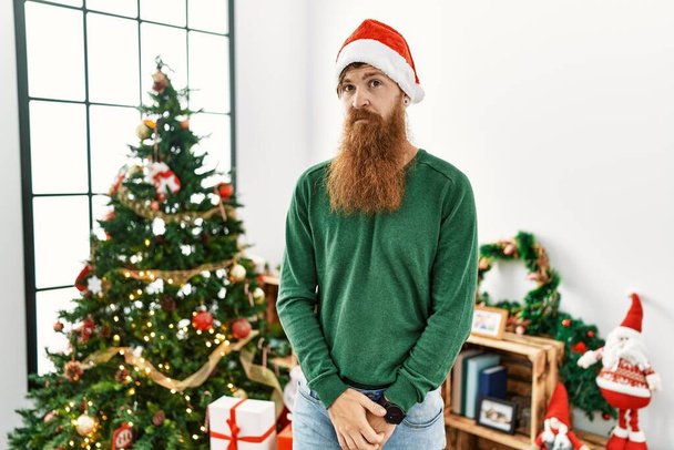 Redhead man with long beard wearing christmas hat by christmas tree relaxed with serious expression on face. simple and natural looking at the camera.  - Photo, Image